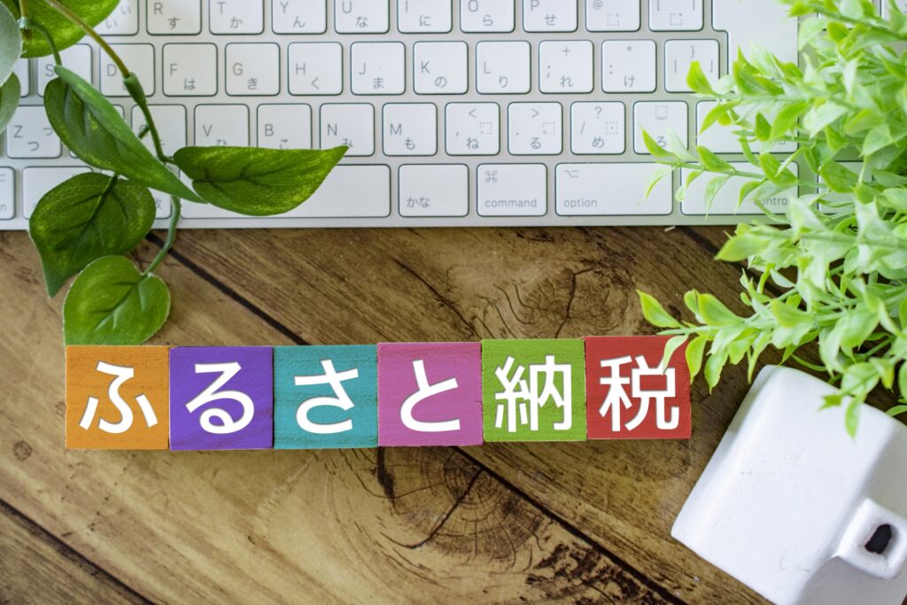 「ふるさと納税」と書かれたカラフルな積み木とキーボード、観葉植物のあるデスク風景